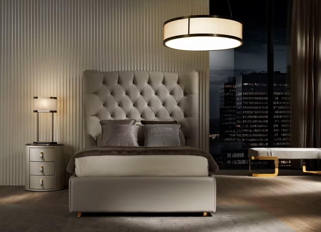 Luxurious bedroom at night with a tufted headboard, neutral bed, and circular ceiling light; city skyline visible through a large window to the right.